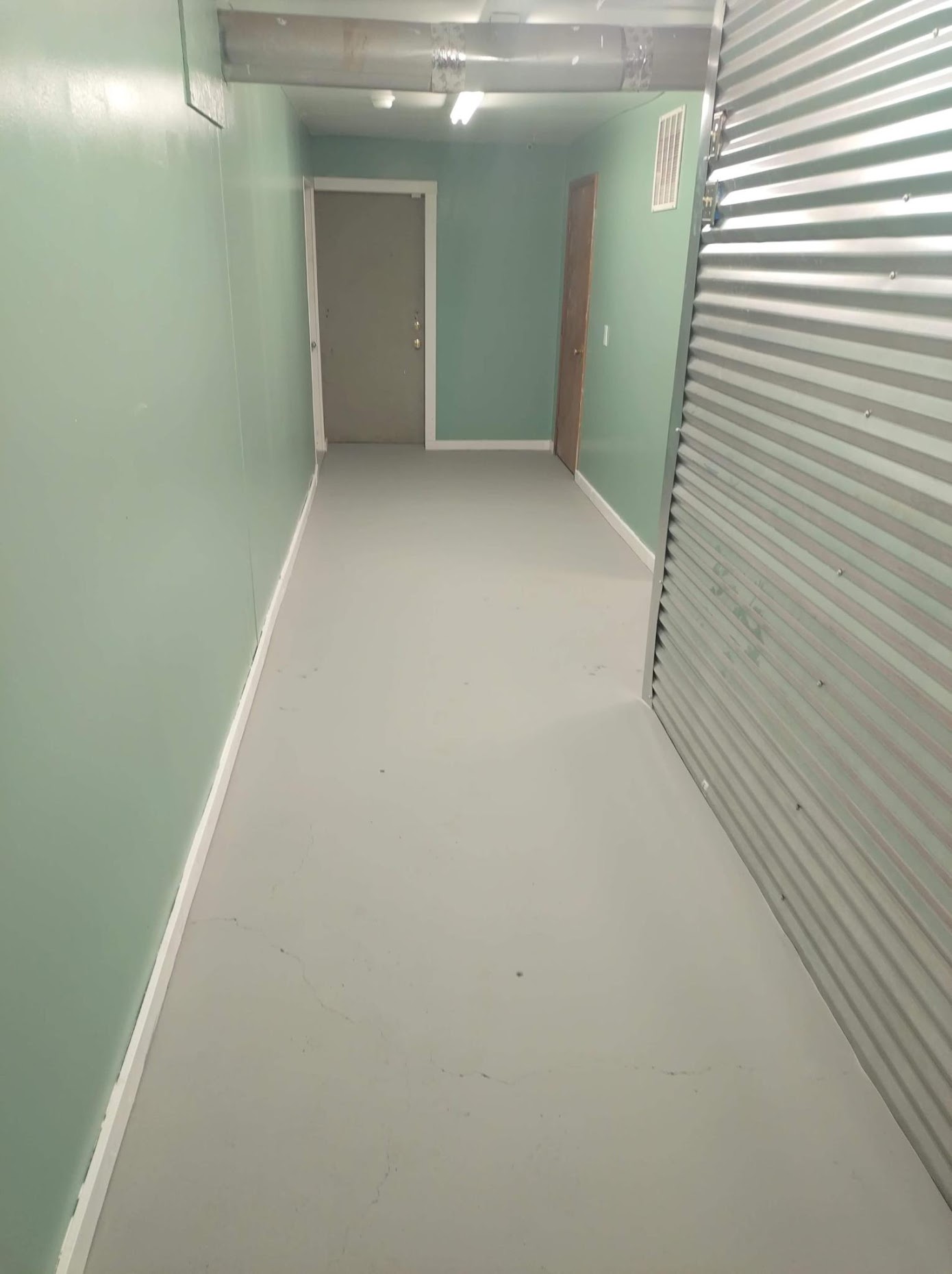 Empty hallway with mint green walls, white trim, fluorescent lighting, and metal corrugated door on right